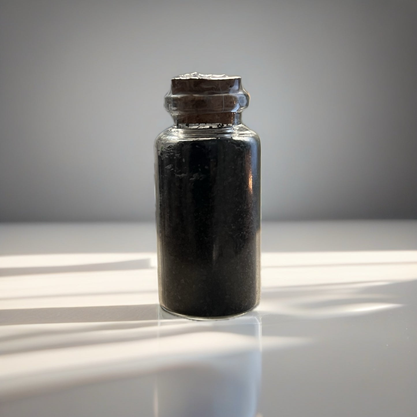 Small glass bottle with cork lid containing a black salt, on a reflective surface.